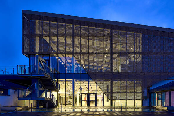 The photograph beautifully encapsulates the contemporary essence and innovative architectural design of the Tabakalera building, the International Centre for Contemporary Culture in San Sebastian, Spain. Shot during the blue hour, the image emphasizes the striking interplay between artificial and natural light, which enhances the modernist aesthetic of the structure.

The building is distinctly characterized by its geometric form, showcasing a sleek, perforated metal facade that creates a semi-transparent veil. This design allows the interior lighting to seep through, giving a dynamic and layered visual texture. The use of glass further accentuates the transparency and openness, inviting viewers to catch glimpses of the interior spaces.

The external staircase featured in the image adds a structural dynamism and serves both a functional and an aesthetic purpose. The sharp lines and precise angles of the staircase contrast with the soft, ambient external lighting, creating a balanced visual composition.

The reflections on the wet ground add an additional layer of depth and intrigue, subtly leading the viewer’s eye into the scene, reinforcing the contemporary and forward-thinking nature of the space.

Overall, the photograph captures the architectural brilliance of the Tabakalera building, highlighting its role not just as a cultural hub but also as an exemplar of modern architectural design.
