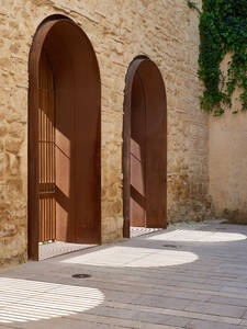 Two arched doorways with rusty metal frames set into a stone wall, with sunlight streaming through the doorway on the left, creating shadows on the ground. The Congress Centre of Cordoba, designed by LAP architects, Spain.